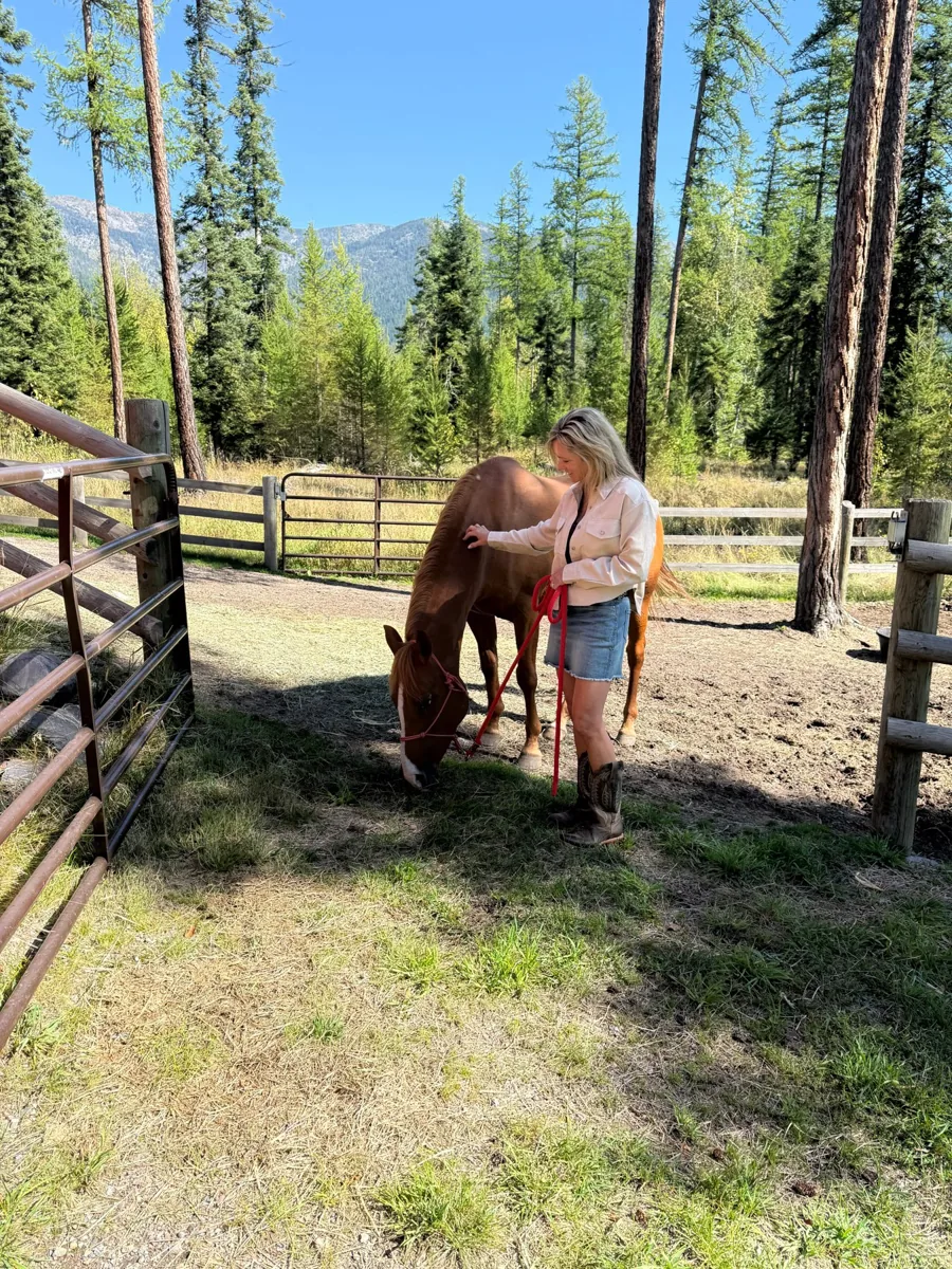 Amy Stevens petting a grazing chestnut horse beneath the Montana mountains